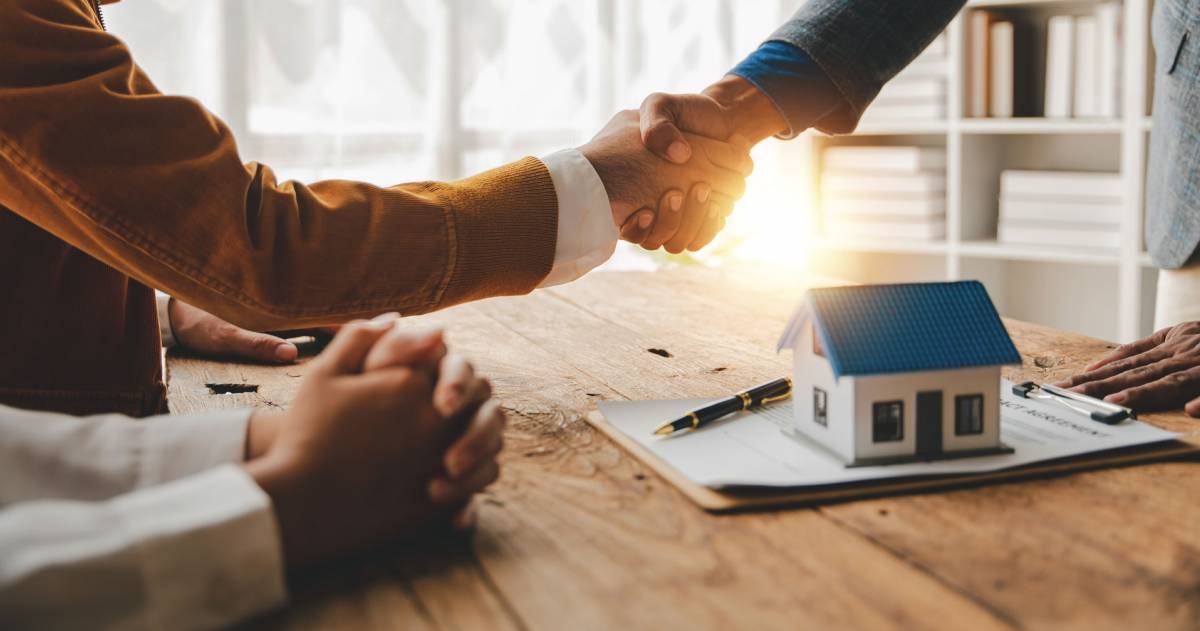 De koopovereenkomst is akkoord. 2 mensen schudden elkaar de hand met op de voorgrond een modelwoning.